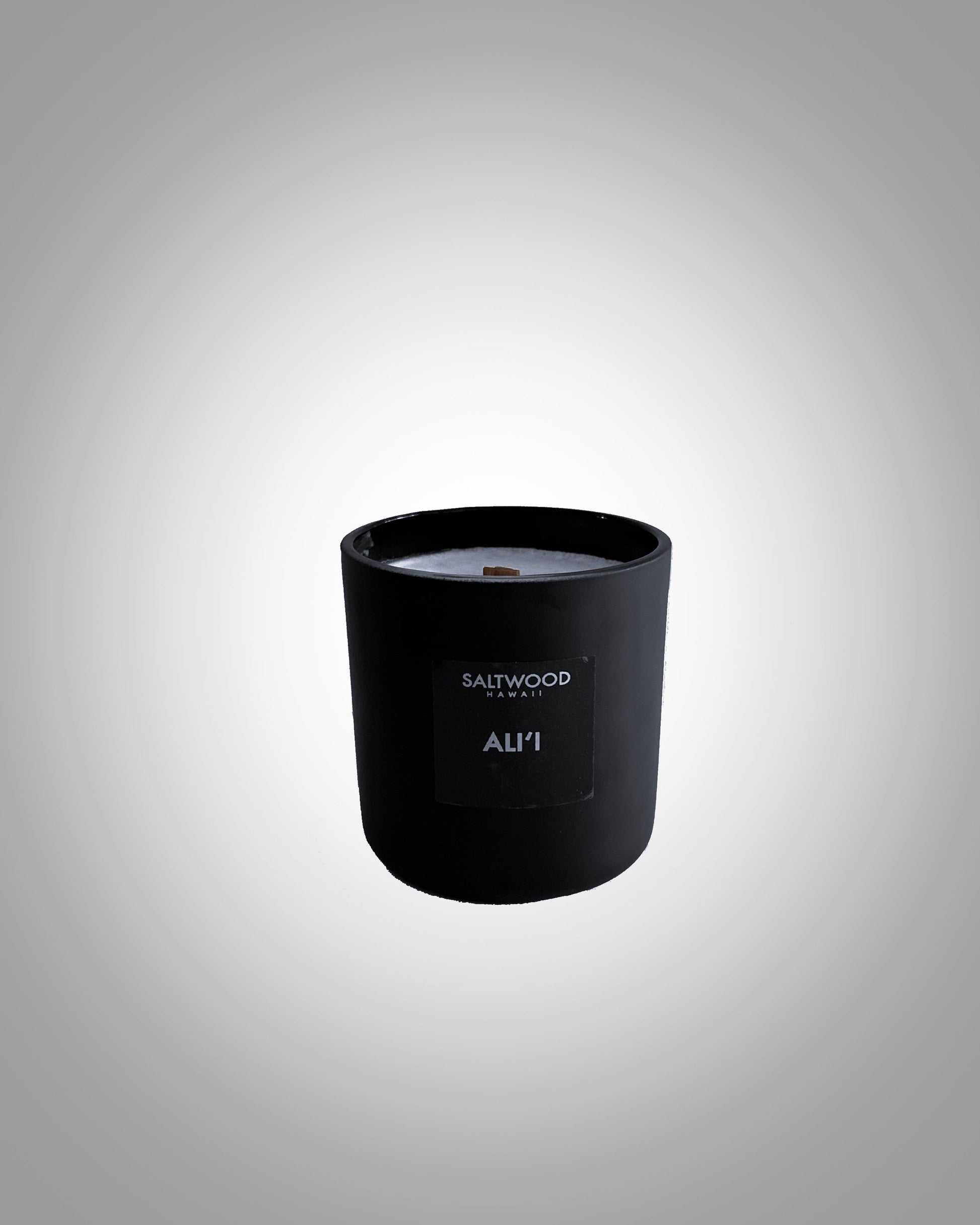 A black candle jar with a white label that reads 'ALI’I' on the front, set against a grey background.